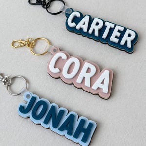 May include: Three custom keychains featuring layered acrylic name tags. The top keychain displays the name CARTER in white on a dark blue base with a black clip. The middle shows CORA in white on a dusty pink base with a gold clip. The bottom shows JONAH in dark blue on a light blue base with a silver clip.