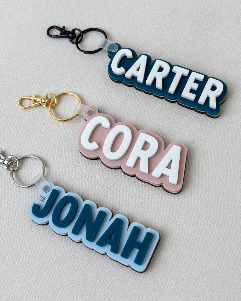 May include: Three custom keychains featuring layered acrylic name tags. The top keychain displays the name CARTER in white on a dark blue base with a black clip. The middle shows CORA in white on a dusty pink base with a gold clip. The bottom shows JONAH in dark blue on a light blue base with a silver clip.