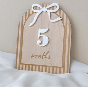 May include: An arched wooden milestone sign featuring a light wood grain finish with tan vertical stripes. The sign displays a raised white number 5 and a white bow at the top, with the word months printed in a tan script font below the number.