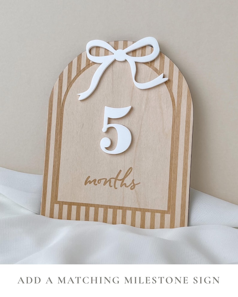 May include: An arched wooden milestone sign featuring a light wood grain finish with tan vertical stripes. The sign displays a raised white number 5 and a white bow at the top, with the word months printed in a tan script font below the number.