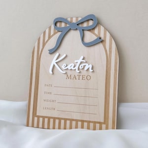 May include: An arched wooden birth announcement sign featuring a light wood grain with vertical tan stripes. A raised gray bow sits at the top. The name Keaton is in white 3D script, with Mateo below in tan. Lines for Date, Time, Weight, and Length are included.