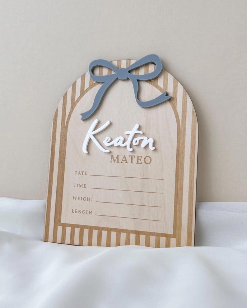 May include: An arched wooden birth announcement sign featuring a light wood grain with vertical tan stripes. A raised gray bow sits at the top. The name Keaton is in white 3D script, with Mateo below in tan. Lines for Date, Time, Weight, and Length are included.