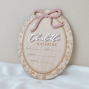 Pu&ograve; includere: Una targa nascita ovale in legno con un fiocco in acrilico rosa polvere sulla parte superiore. Al centro il nome Charlotte in rilievo bianco, seguito da Katarine e linee per data, ora, peso e lunghezza. Il bordo presenta un motivo floreale.