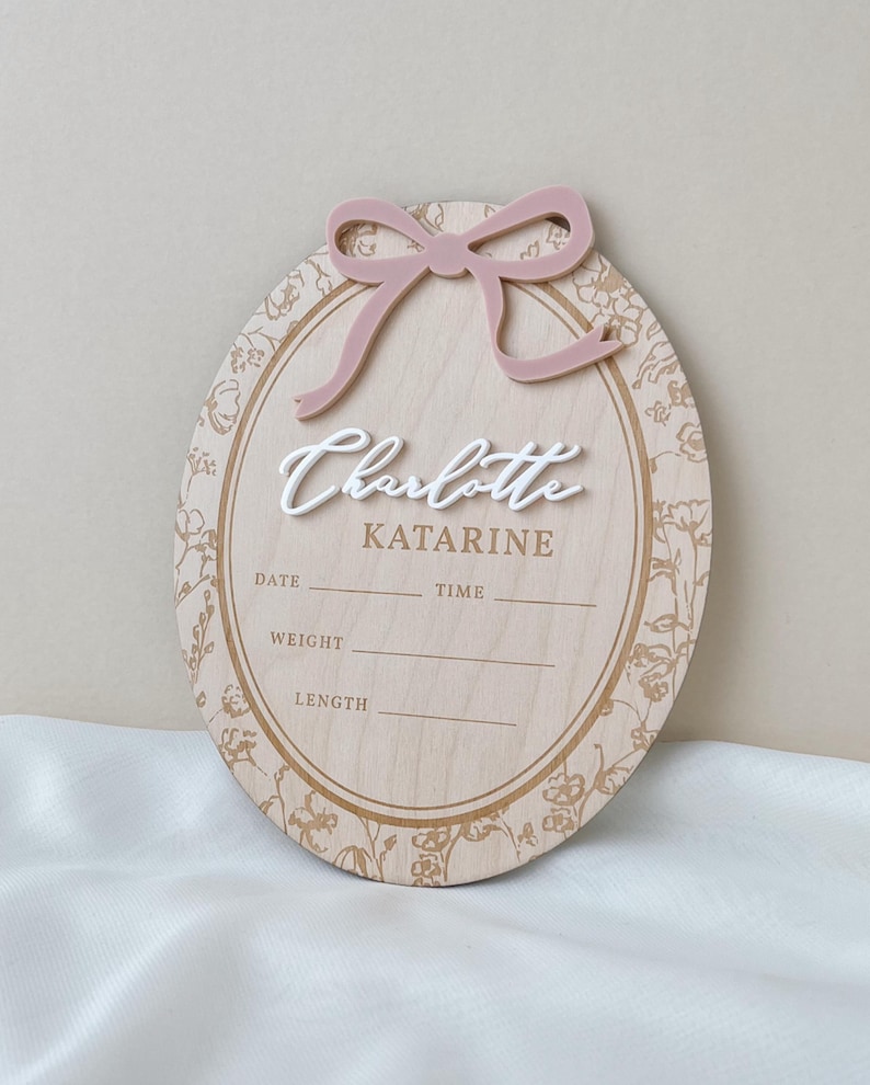 Pu&ograve; includere: Una targa nascita ovale in legno con un fiocco in acrilico rosa polvere sulla parte superiore. Al centro il nome Charlotte in rilievo bianco, seguito da Katarine e linee per data, ora, peso e lunghezza. Il bordo presenta un motivo floreale.