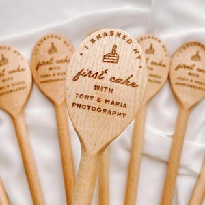 K&ouml;nnte beinhalten: Eine Sammlung von hellen Holzl&ouml;ffeln, die jeweils ein lasergraviertes Design eines kleinen Kuchensymbols und den Text "I smashed my first cake with Tony & Maria Photography" mittig auf der L&ouml;ffellaffe in klarer Schrift zeigen.