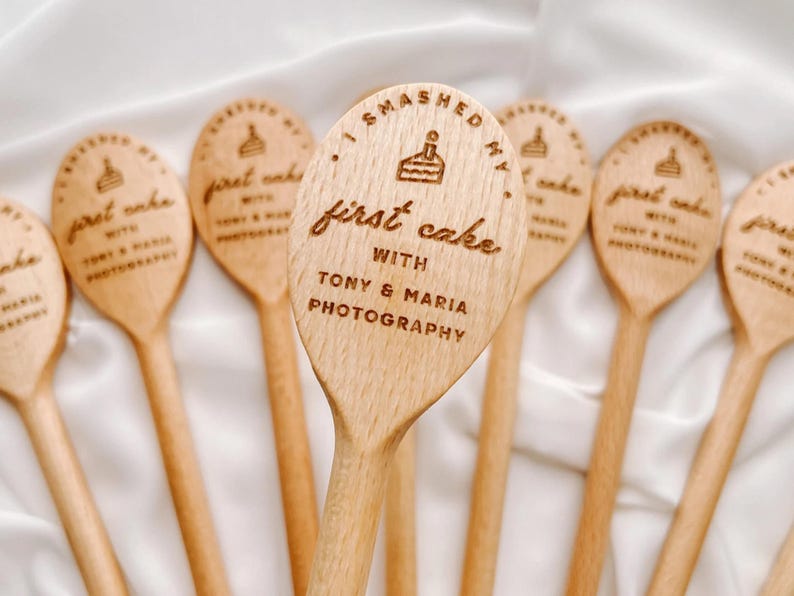 K&ouml;nnte beinhalten: Eine Sammlung von hellen Holzl&ouml;ffeln, die jeweils ein lasergraviertes Design eines kleinen Kuchensymbols und den Text "I smashed my first cake with Tony & Maria Photography" mittig auf der L&ouml;ffellaffe in klarer Schrift zeigen.