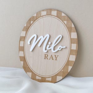 K&ouml;nnte beinhalten: Ein ovales Holzschild mit einem beigen und hellbraunen Karomuster am Rand. In der Mitte steht der Name Milo in erhabener, wei&szlig;er Schreibschrift, darunter ist der Name RAY in einer einfachen braunen serifenlosen Schrift aufgedruckt.