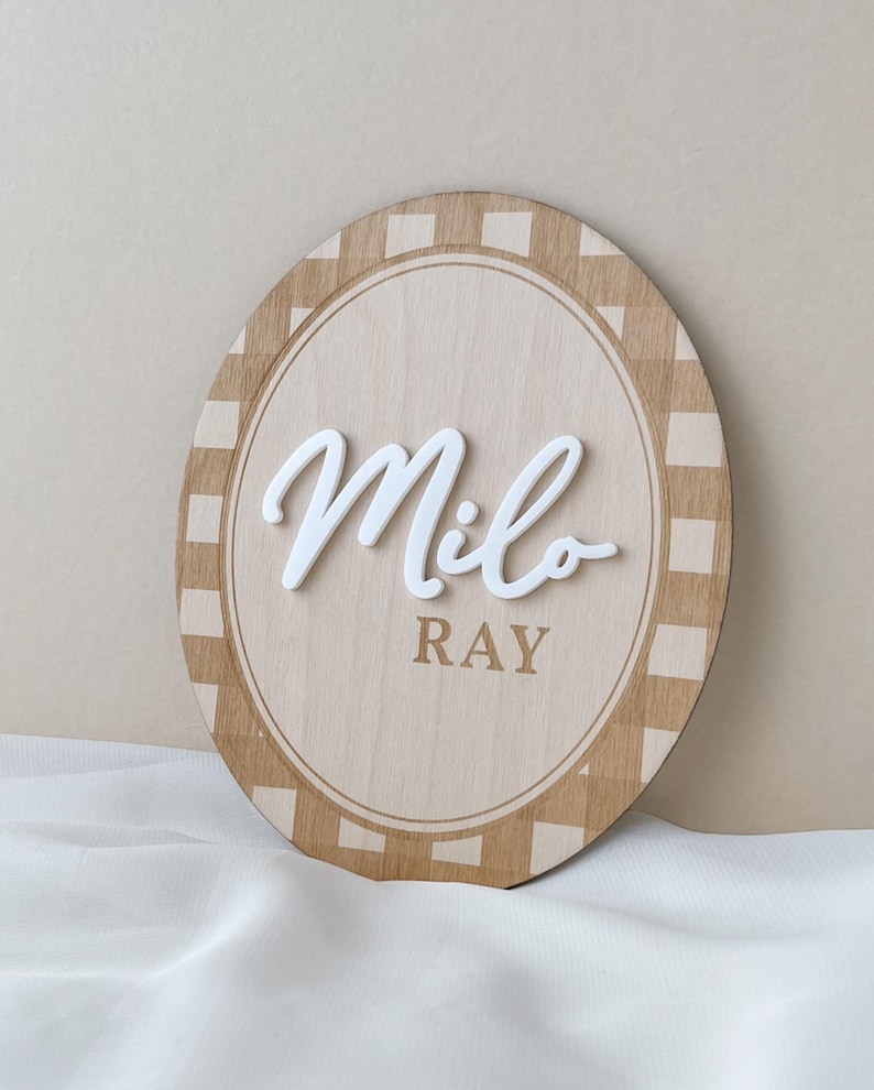 K&ouml;nnte beinhalten: Ein ovales Holzschild mit einem beigen und hellbraunen Karomuster am Rand. In der Mitte steht der Name Milo in erhabener, wei&szlig;er Schreibschrift, darunter ist der Name RAY in einer einfachen braunen serifenlosen Schrift aufgedruckt.