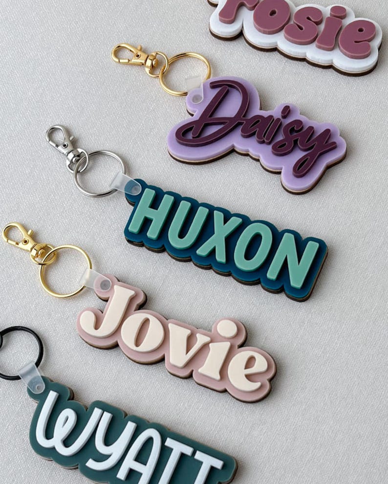 May include: A collection of layered acrylic keychains featuring custom names in various colors including purple, teal, beige, and white. Each keychain has a metal lobster clasp and ring attached to a multi-layered, scalloped-edge name plate.