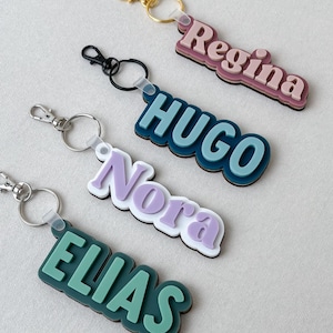 May include: Four custom keychains featuring raised, layered lettering in pastel and bold colors. The names Regina, HUGO, Nora, and ELIAS are displayed in distinct fonts and color combinations, each attached to a metal split ring with a lobster clasp.