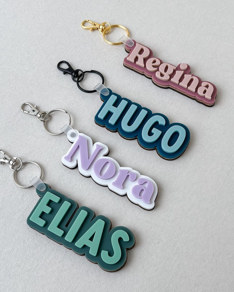 May include: Four custom keychains featuring raised, layered lettering in pastel and bold colors. The names Regina, HUGO, Nora, and ELIAS are displayed in distinct fonts and color combinations, each attached to a metal split ring with a lobster clasp.