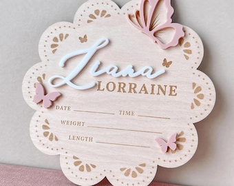 Op de afbeelding: Een lichtroze houten geboorteaankondigingsbord in de vorm van een geschulpte bloem. De naam "Liana Lorraine" wordt weergegeven in witte letters. Roze vlinders en decoratieve uitsparingen sieren de plaat, met ruimtes voor datum, tijd, gewicht en lengte.