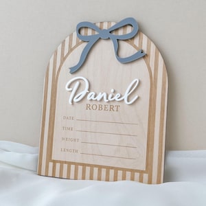 May include: An arched wooden birth announcement sign featuring a light wood grain with vertical tan stripes. A raised blue bow sits at the top. The name Daniel Robert is displayed in white and tan text, with lines for date, time, weight, and length.