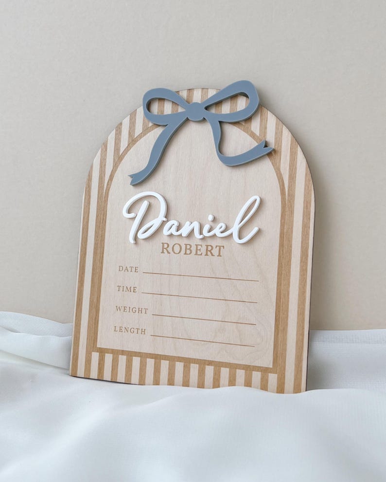 May include: An arched wooden birth announcement sign featuring a light wood grain with vertical tan stripes. A raised blue bow sits at the top. The name Daniel Robert is displayed in white and tan text, with lines for date, time, weight, and length.
