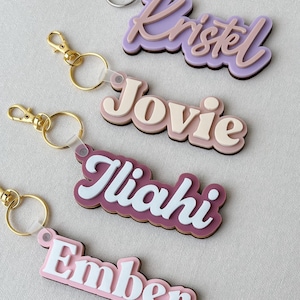 May include: Four custom name keychains featuring layered acrylic designs. The keychains display the names Kristel, Jovie, Iliahi, and Ember in various pastel and bold colors, each attached to a metal split ring and a lobster claw clasp.