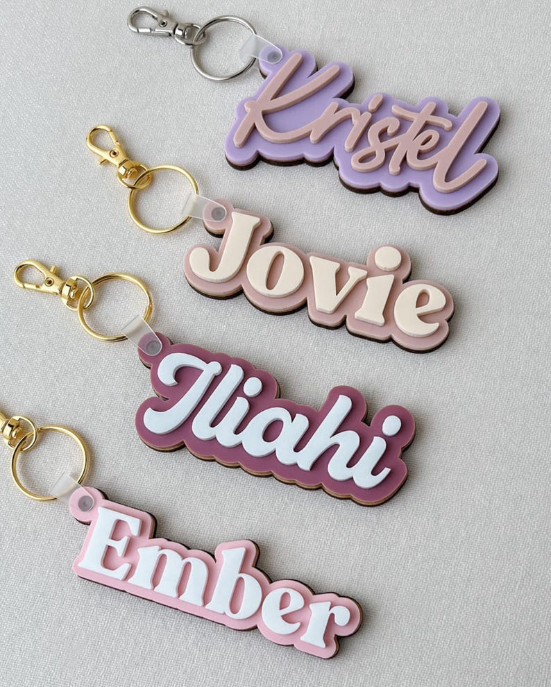 May include: Four custom name keychains featuring layered acrylic designs. The keychains display the names Kristel, Jovie, Iliahi, and Ember in various pastel and bold colors, each attached to a metal split ring and a lobster claw clasp.