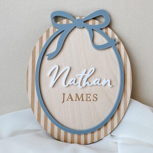 Puede incluir: Un letrero de madera ovalado con un lazo y borde azul claro. El letrero tiene rayas verticales y el nombre "Nathan" en escritura blanca sobre "JAMES" en una fuente serif. Un art&iacute;culo decorativo.