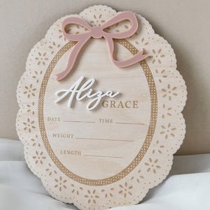 May include: An oval wooden birth announcement sign with a scalloped edge featuring laser-cut floral patterns. It includes a dusty pink acrylic bow at the top and the white 3D script name Aliza Grace. Printed lines provide spaces for date, time, weight, and length.
