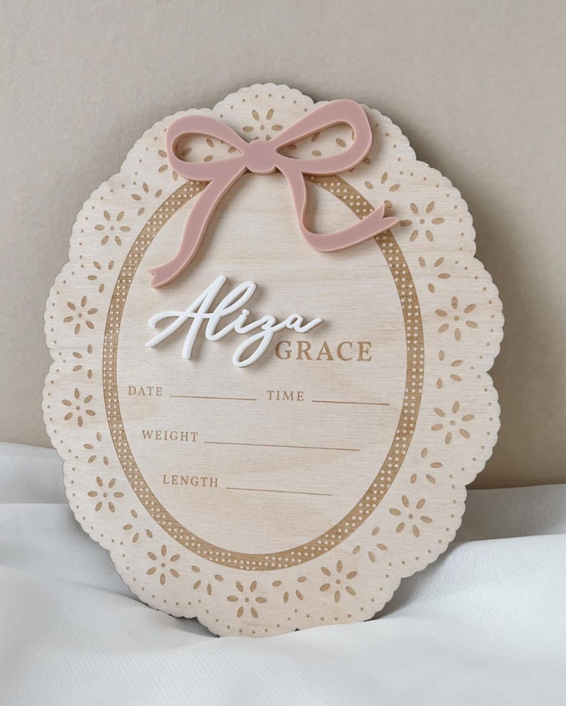 May include: An oval wooden birth announcement sign with a scalloped edge featuring laser-cut floral patterns. It includes a dusty pink acrylic bow at the top and the white 3D script name Aliza Grace. Printed lines provide spaces for date, time, weight, and length.