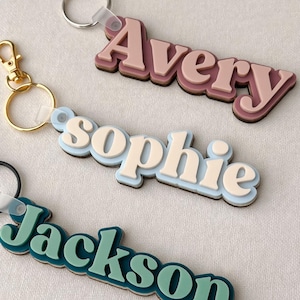 Puede incluir: Tres llaveros personalizados con nombres en un dise&ntilde;o en capas. Los nombres "Avery", "Sophie" y "Jackson" se muestran en una fuente retro. Cada llavero tiene una combinaci&oacute;n de colores diferente, incluyendo rosa, azul y verde azulado.