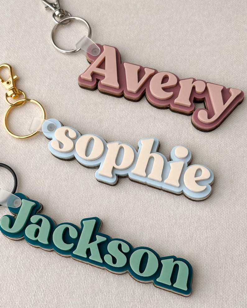 Puede incluir: Tres llaveros personalizados con nombres en un dise&ntilde;o en capas. Los nombres "Avery", "Sophie" y "Jackson" se muestran en una fuente retro. Cada llavero tiene una combinaci&oacute;n de colores diferente, incluyendo rosa, azul y verde azulado.