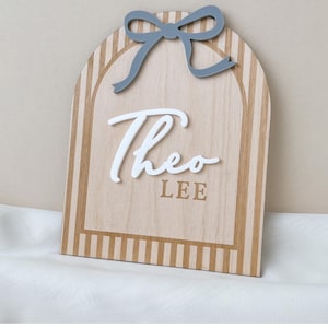May include: An arched wooden sign featuring a light wood grain finish with vertical tan stripes. A raised white script name Theo is centered above the block text LEE. A solid grey bow is attached to the top center of the arched wooden plaque.