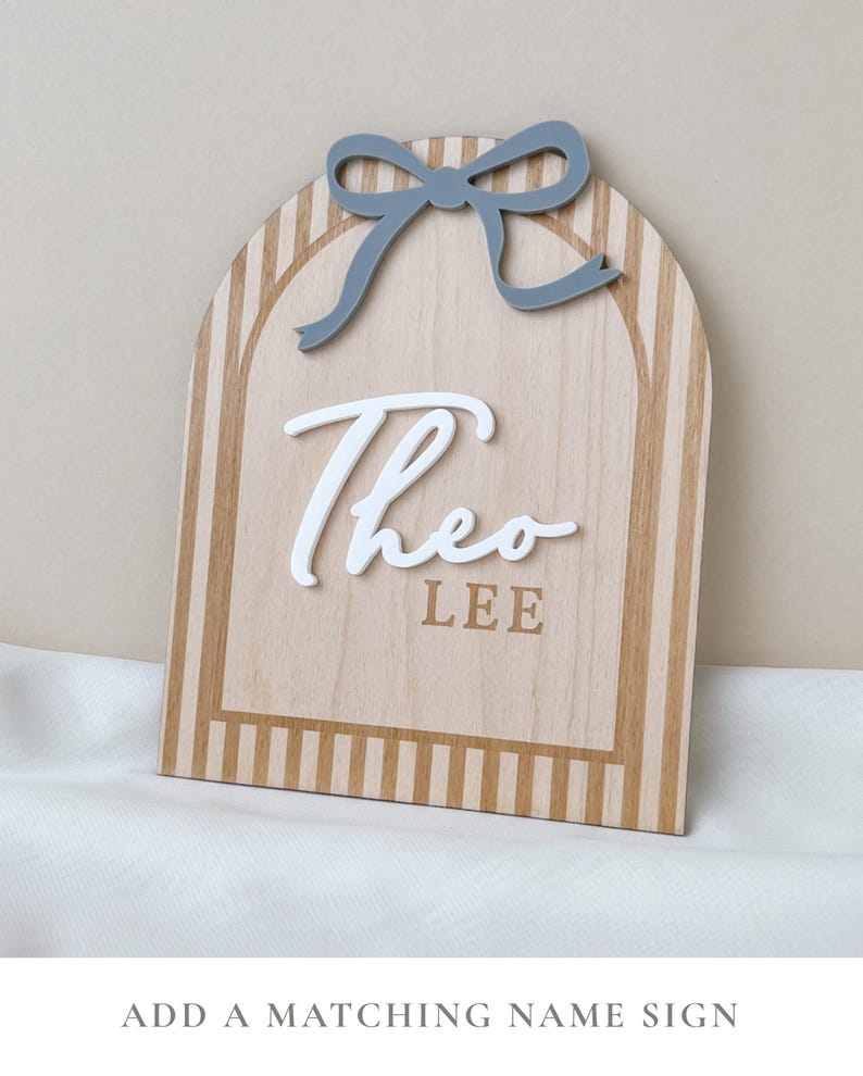 May include: An arched wooden sign featuring a light wood grain finish with vertical tan stripes. A raised white script name Theo is centered above the block text LEE. A solid grey bow is attached to the top center of the arched wooden plaque.