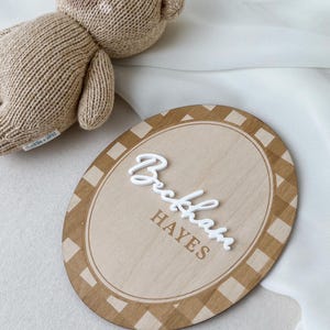 K&ouml;nnte beinhalten: Ein beigefarbener, gestrickter Teddyb&auml;r mit einem kleinen wei&szlig;en Etikett und ein ovales Holzschild mit einem braun-wei&szlig;en Karomuster am Rand. Das Schild zeigt den Namen Beckham in erhabener, wei&szlig;er Schreibschrift und HAYES in braunen Druckbuchstaben.