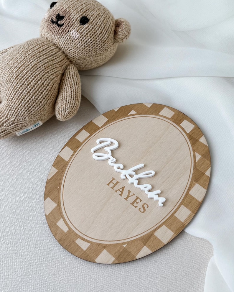 K&ouml;nnte beinhalten: Ein beigefarbener, gestrickter Teddyb&auml;r mit einem kleinen wei&szlig;en Etikett und ein ovales Holzschild mit einem braun-wei&szlig;en Karomuster am Rand. Das Schild zeigt den Namen Beckham in erhabener, wei&szlig;er Schreibschrift und HAYES in braunen Druckbuchstaben.