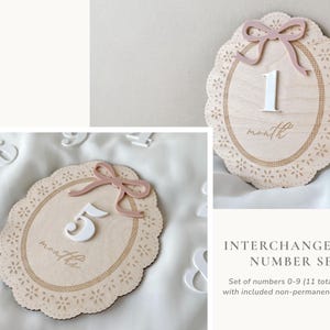 May include: An oval wooden milestone plaque with a scalloped edge, a dusty pink bow, and a white number. The plaque features laser-cut floral details and the word month or months. The set includes interchangeable white numbers from zero to nine.