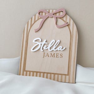 May include: An arched wooden sign featuring a light wood grain finish with vertical stripes. A dusty pink acrylic bow is attached to the top. The name Stella is written in raised white script lettering, with the name JAMES printed in brown below it.