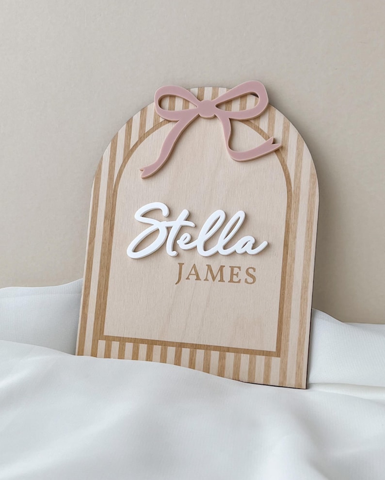 May include: An arched wooden sign featuring a light wood grain finish with vertical stripes. A dusty pink acrylic bow is attached to the top. The name Stella is written in raised white script lettering, with the name JAMES printed in brown below it.