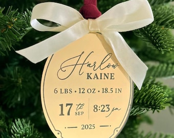 May include: An oval gold-toned acrylic ornament featuring the name Harlow Kaine, birth details 6 lbs 12 oz and 18.5 in, the date 17th Sep 2025, and the time 8:23 pm. The ornament is topped with a cream-colored satin ribbon bow and a dark red velvet loop.