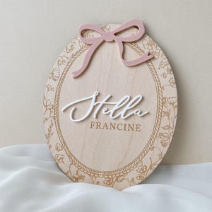 Pu&ograve; includere: Una targa ovale in legno con finitura a venature chiare e bordo floreale dorato. Un fiocco rosa polvere in rilievo &egrave; fissato nella parte superiore. Il nome Stella &egrave; scritto in corsivo bianco 3D, con il nome FRANCINE stampato sotto in oro.