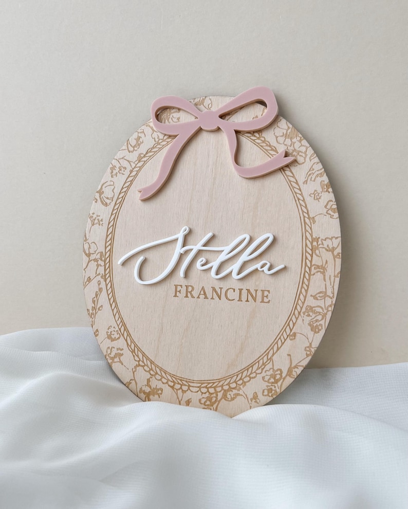 Pu&ograve; includere: Una targa ovale in legno con finitura a venature chiare e bordo floreale dorato. Un fiocco rosa polvere in rilievo &egrave; fissato nella parte superiore. Il nome Stella &egrave; scritto in corsivo bianco 3D, con il nome FRANCINE stampato sotto in oro.