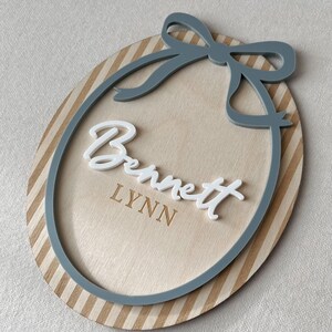 Puede incluir: Letrero ovalado de madera con borde a rayas y un lazo gris. El letrero presenta el nombre "Bennett" en escritura blanca y el nombre "Lynn" en una fuente m&aacute;s peque&ntilde;a y grabada. El dise&ntilde;o es en capas.