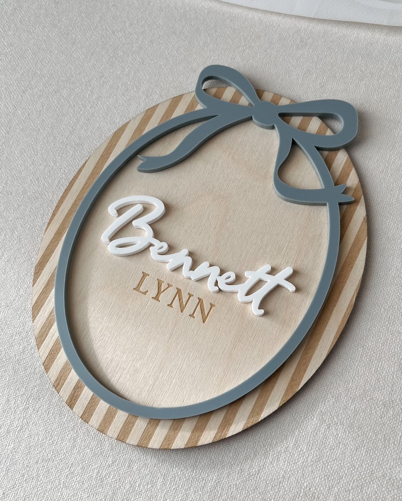 Puede incluir: Letrero ovalado de madera con borde a rayas y un lazo gris. El letrero presenta el nombre "Bennett" en escritura blanca y el nombre "Lynn" en una fuente m&aacute;s peque&ntilde;a y grabada. El dise&ntilde;o es en capas.