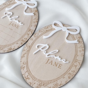 Pu&ograve; includere: Due targhe ovali in legno con finitura in legno chiaro, bordo floreale stampato e decorazione a corda. Ogni targa presenta un fiocco bianco in 3D sulla parte superiore e il nome Paloma JANE in corsivo bianco. Una targa riporta la parola TIME.