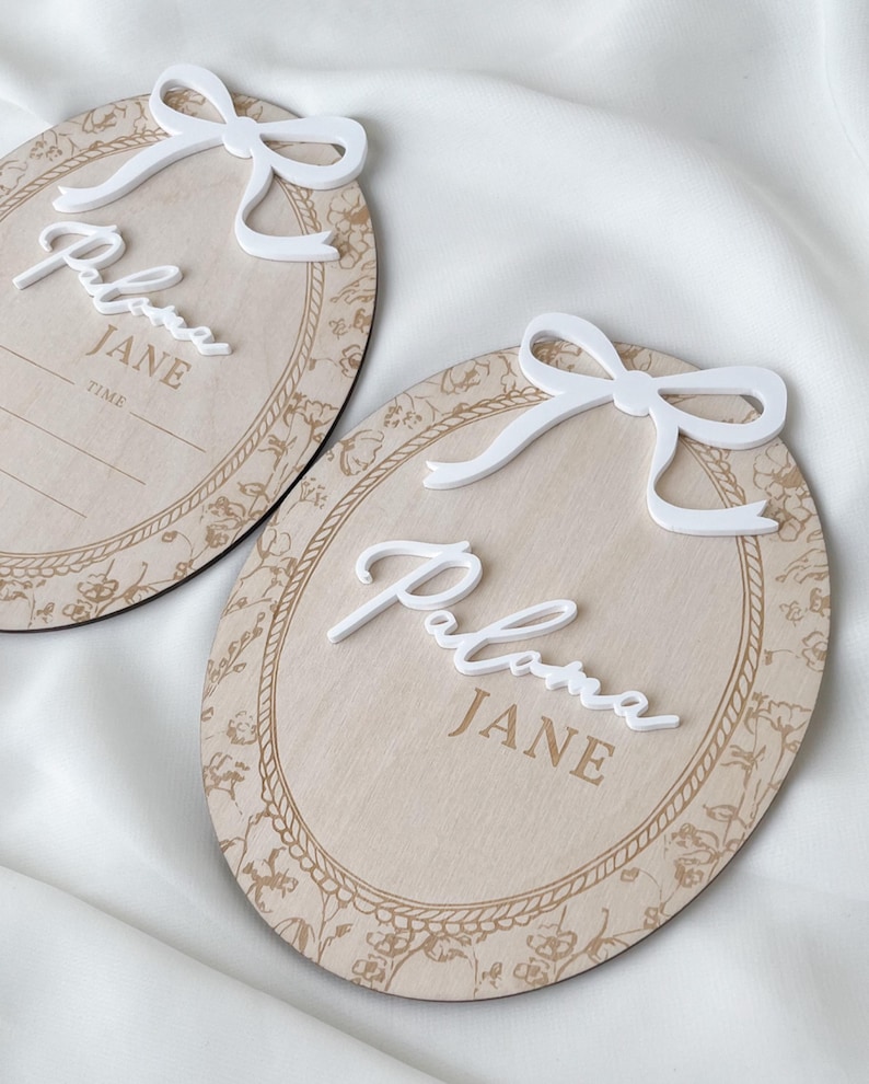 Pu&ograve; includere: Due targhe ovali in legno con finitura in legno chiaro, bordo floreale stampato e decorazione a corda. Ogni targa presenta un fiocco bianco in 3D sulla parte superiore e il nome Paloma JANE in corsivo bianco. Una targa riporta la parola TIME.