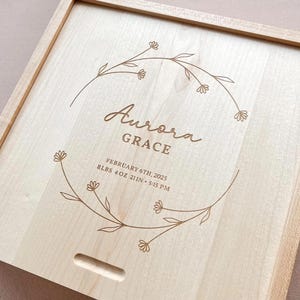 May include: A square light-coloured wooden box with a sliding lid featuring a laser-engraved floral wreath design. The text reads Aurora GRACE, FEBRUARY 6TH, 2025, 8LBS 4OZ 21IN 5:15 PM. The box has a small rectangular cutout for sliding the lid.