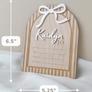 May include: An arched wooden birth announcement sign featuring a white 3D bow, the name Kaidyn Jo, and lines for date, time, weight, and length. The sign has a light wood grain with vertical stripes and measures 6.5 inches by 5.25 inches.