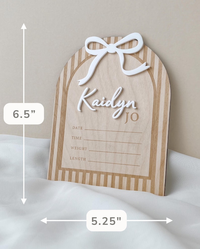 May include: An arched wooden birth announcement sign featuring a white 3D bow, the name Kaidyn Jo, and lines for date, time, weight, and length. The sign has a light wood grain with vertical stripes and measures 6.5 inches by 5.25 inches.