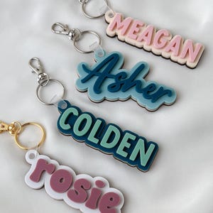 May include: Four custom keychains featuring layered acrylic name tags in various colors and fonts. The keychains include metal lobster claw clasps and split rings. The names displayed are MEAGAN in pink, Asher in blue, COLDEN in green, and rosie in mauve.