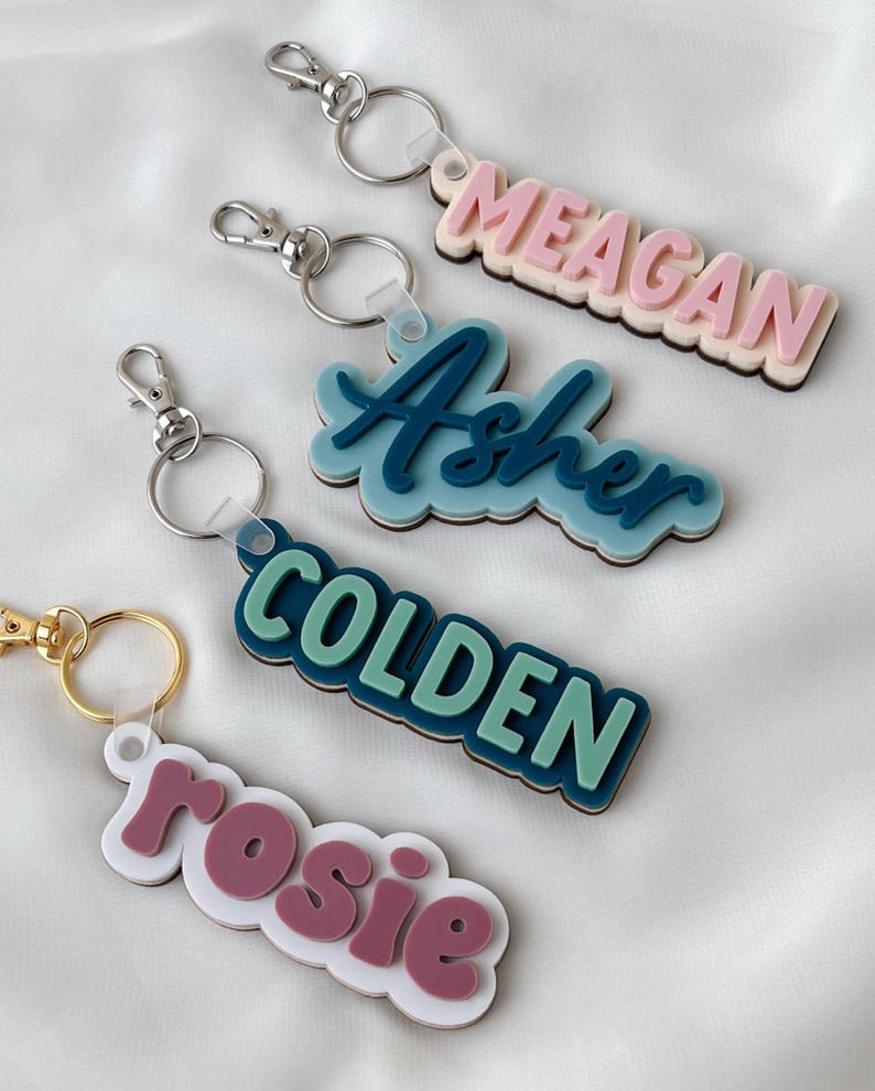 May include: Four custom keychains featuring layered acrylic name tags in various colors and fonts. The keychains include metal lobster claw clasps and split rings. The names displayed are MEAGAN in pink, Asher in blue, COLDEN in green, and rosie in mauve.