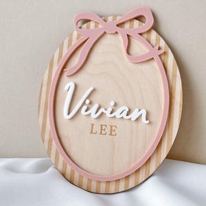 Puede incluir: Letrero ovalado de madera con lazo y borde rosa. El letrero presenta el nombre "Vivian" en escritura blanca, con "LEE" debajo. El fondo tiene rayas verticales en color marr&oacute;n claro.