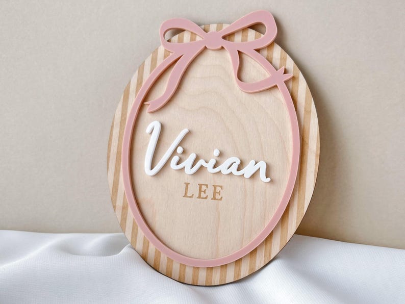 Puede incluir: Letrero ovalado de madera con lazo y borde rosa. El letrero presenta el nombre "Vivian" en escritura blanca, con "LEE" debajo. El fondo tiene rayas verticales en color marr&oacute;n claro.
