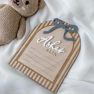 May include: A light brown knitted teddy bear next to an arched wooden birth announcement sign featuring a blue acrylic bow, the white 3D name Asher, the brown text ALIN, and printed lines for date, time, weight, and length information.