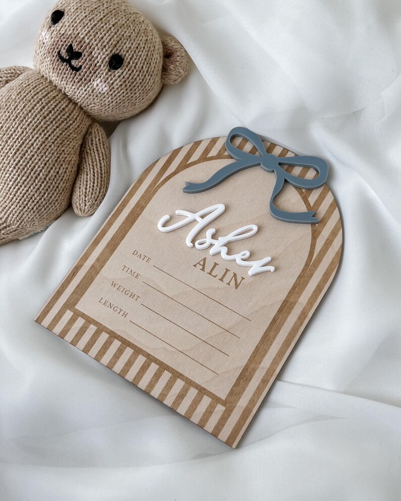 May include: A light brown knitted teddy bear next to an arched wooden birth announcement sign featuring a blue acrylic bow, the white 3D name Asher, the brown text ALIN, and printed lines for date, time, weight, and length information.