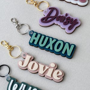 May include: A collection of five custom keychains featuring layered, multi-colored acrylic name tags. Each keychain displays a unique name including Rosie, Daisy, HUXON, Jovie, and WYATT, attached to a gold or silver metal split ring and lobster clasp.