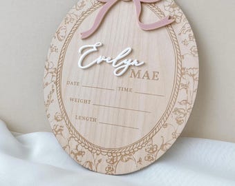 May include: An oval wooden birth announcement sign featuring a dusty pink acrylic bow at the top. The name Evelyn Mae is displayed in white script and serif lettering. The board includes engraved fields for date, time, weight, and length.